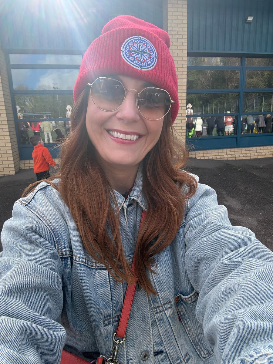 A red ribbed knit beanie hat with a folded cuff, featuring a circular embroidered compass design in blue, white, and red on the front centre. Keep your warm and comfy on your confidence recovery journey. Red Front angel with model